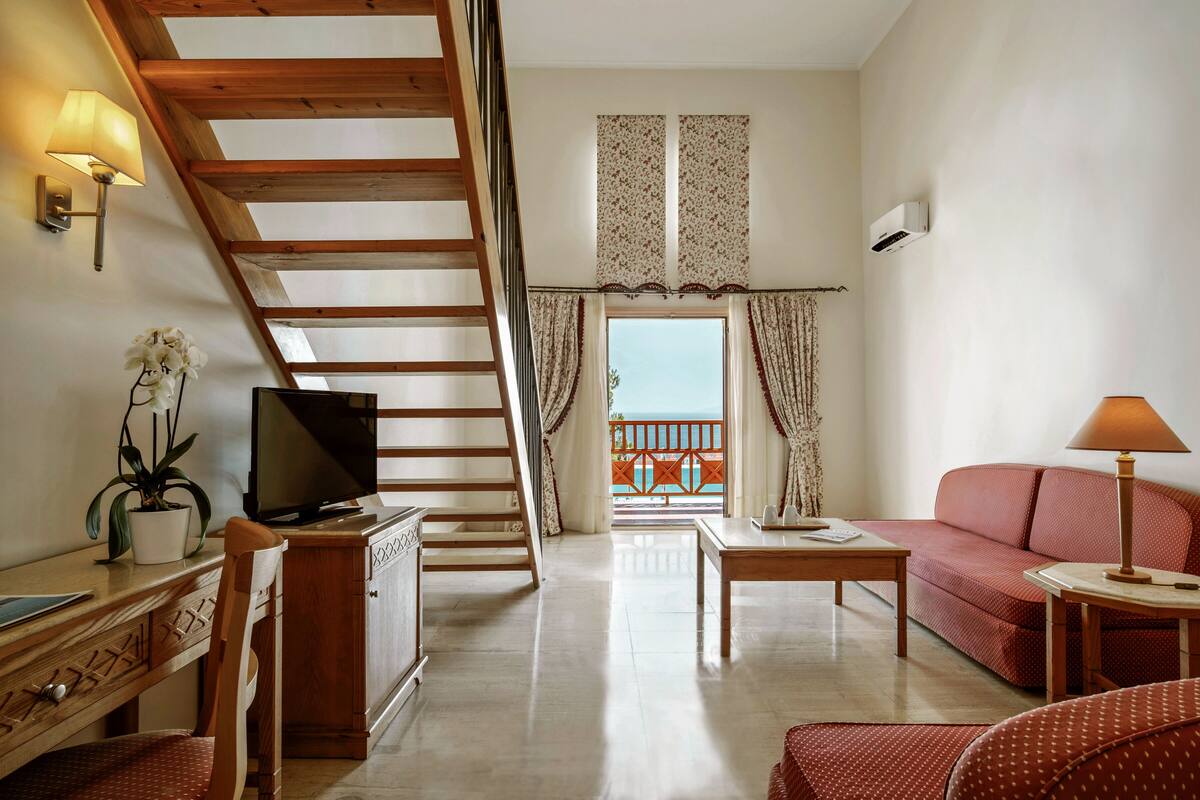 A living room with a wooden staircase, a television, a sofa, a coffee table, and a balcony with curtains.