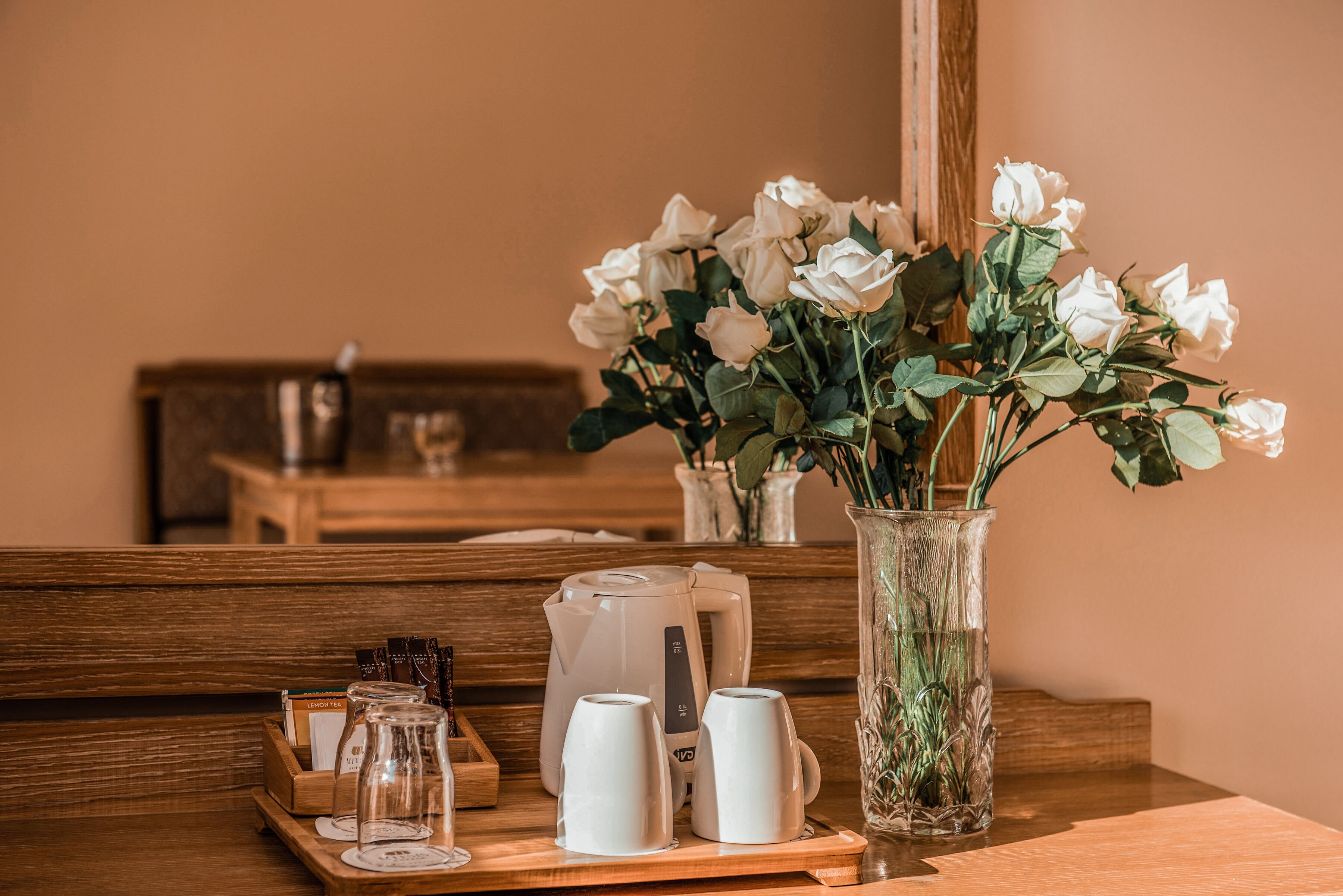 Una mesa de madera con una bandeja que contiene una cafetera, dos tazas y un tarro de vidrio. También se observa un florero con rosas blancas.