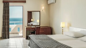 A hotel room with a bed, a desk with a chair, a large mirror, a nightstand with a lamp, and a view of the sea through a sliding door. - Canvas by Mitsis Family Village (Kos)