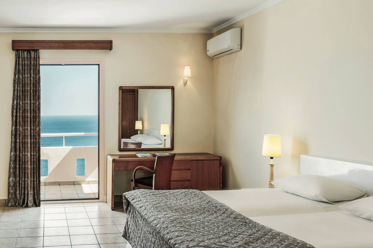 A hotel room with a bed, a desk with a chair, a large mirror, a nightstand with a lamp, and a view of the sea through a sliding door.