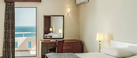 A hotel room with a bed, a desk with a chair, a large mirror, a nightstand with a lamp, and a view of the sea through a sliding door.