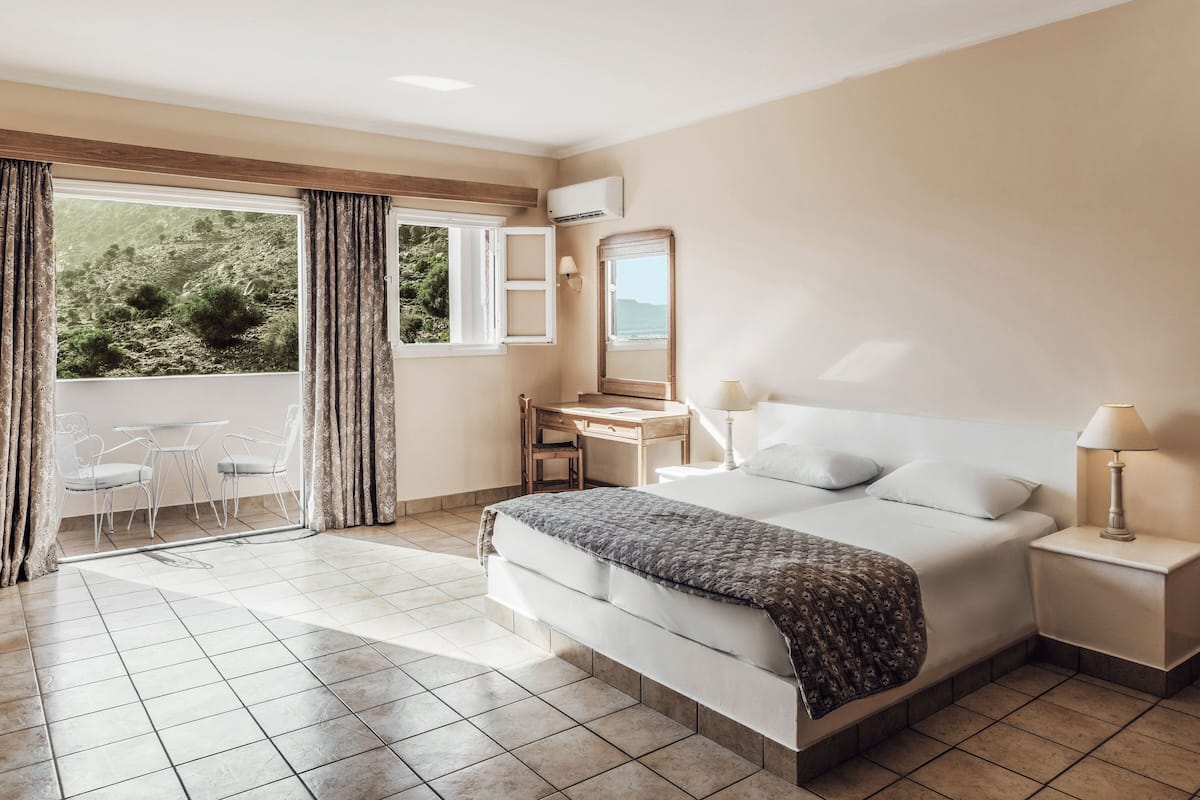 A hotel room with a bed, bedside table, desk, chair, and a view of greenery through the window.
