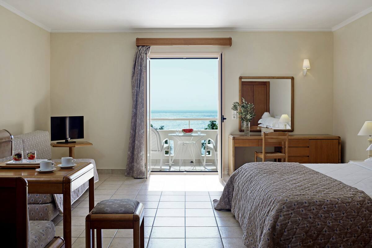 A hotel room with a bed, a desk, a television, and a balcony with a view of the sea.