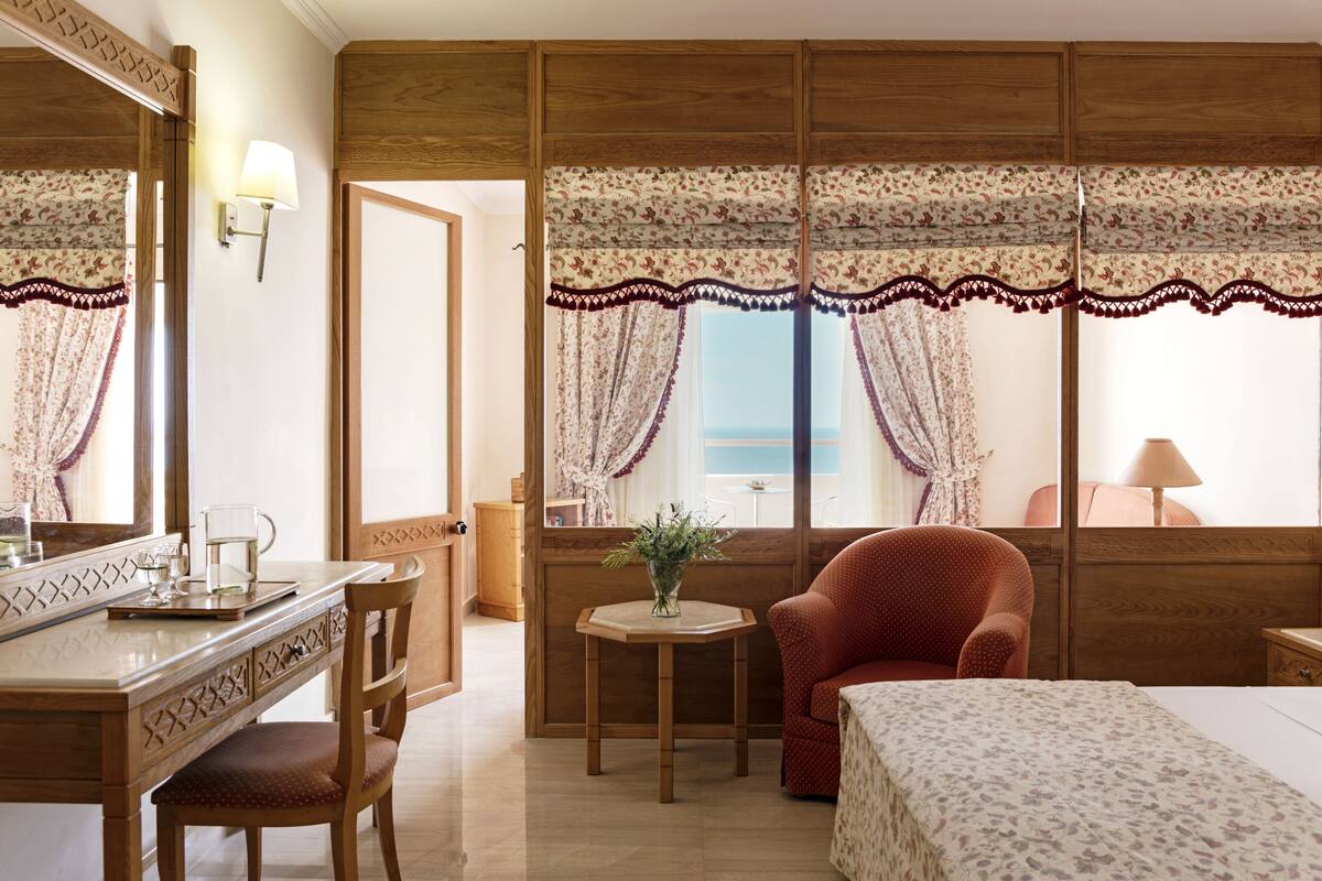 A hotel room with a wooden wardrobe, a desk with a mirror, a red armchair, a small round table with a vase, and a bed with a floral bedspread.