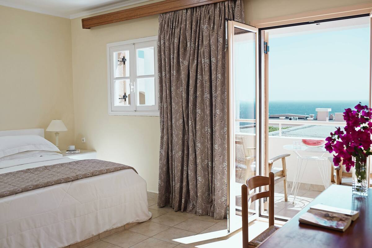 A hotel room with a bed, a table with a chair, a window with curtains, and a view of the sea.