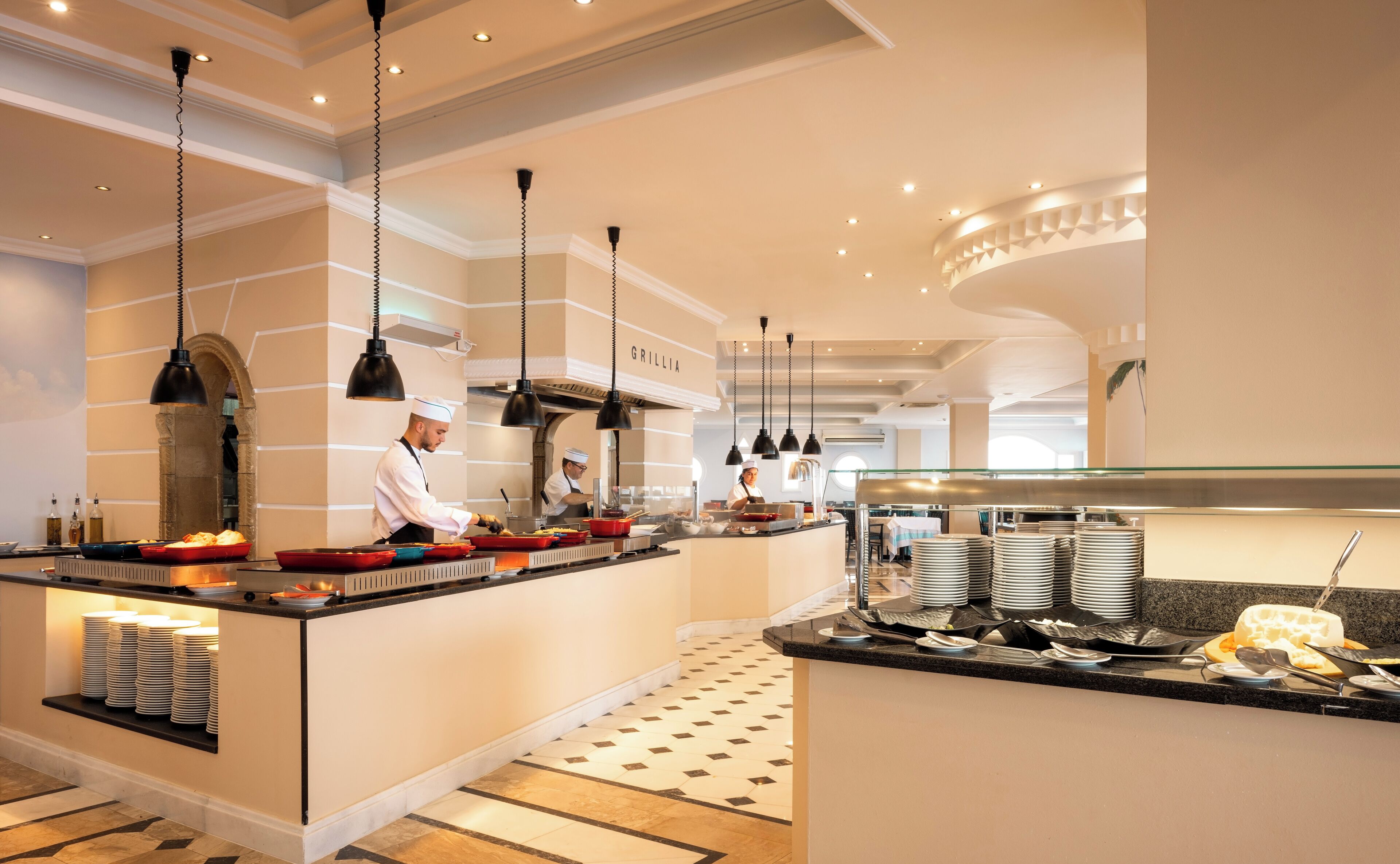 A modern kitchen with chefs preparing food, a buffet area with plates and bowls, and a clean, well-lit interior.