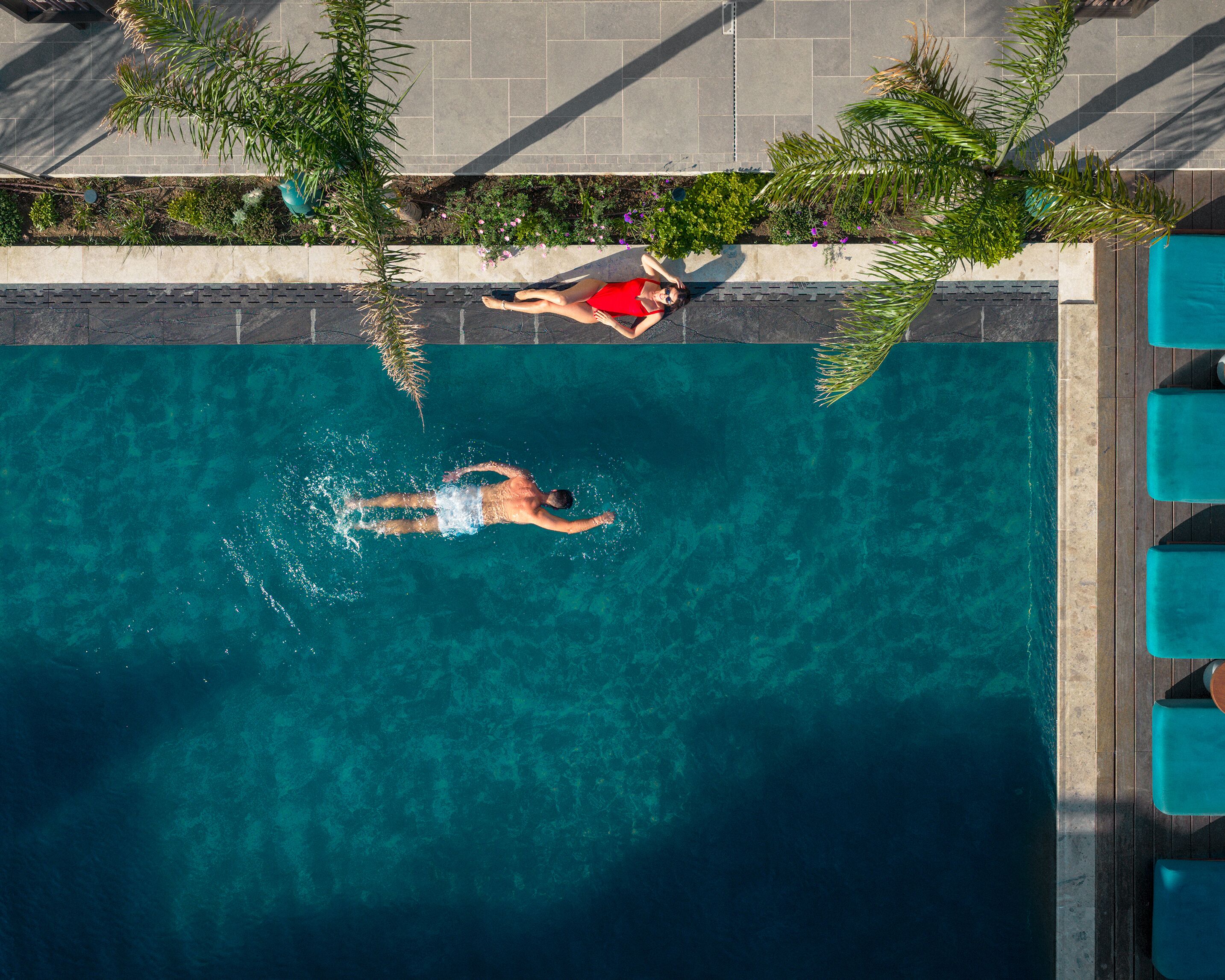 Una piscina azul cristalina con una persona nadando, rodeada de un deck con baldosas y vegetación.