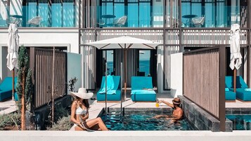 Zona de piscina con sillones de descanso y sombrillas, vista al océano y un edificio moderno con grandes ventanales.