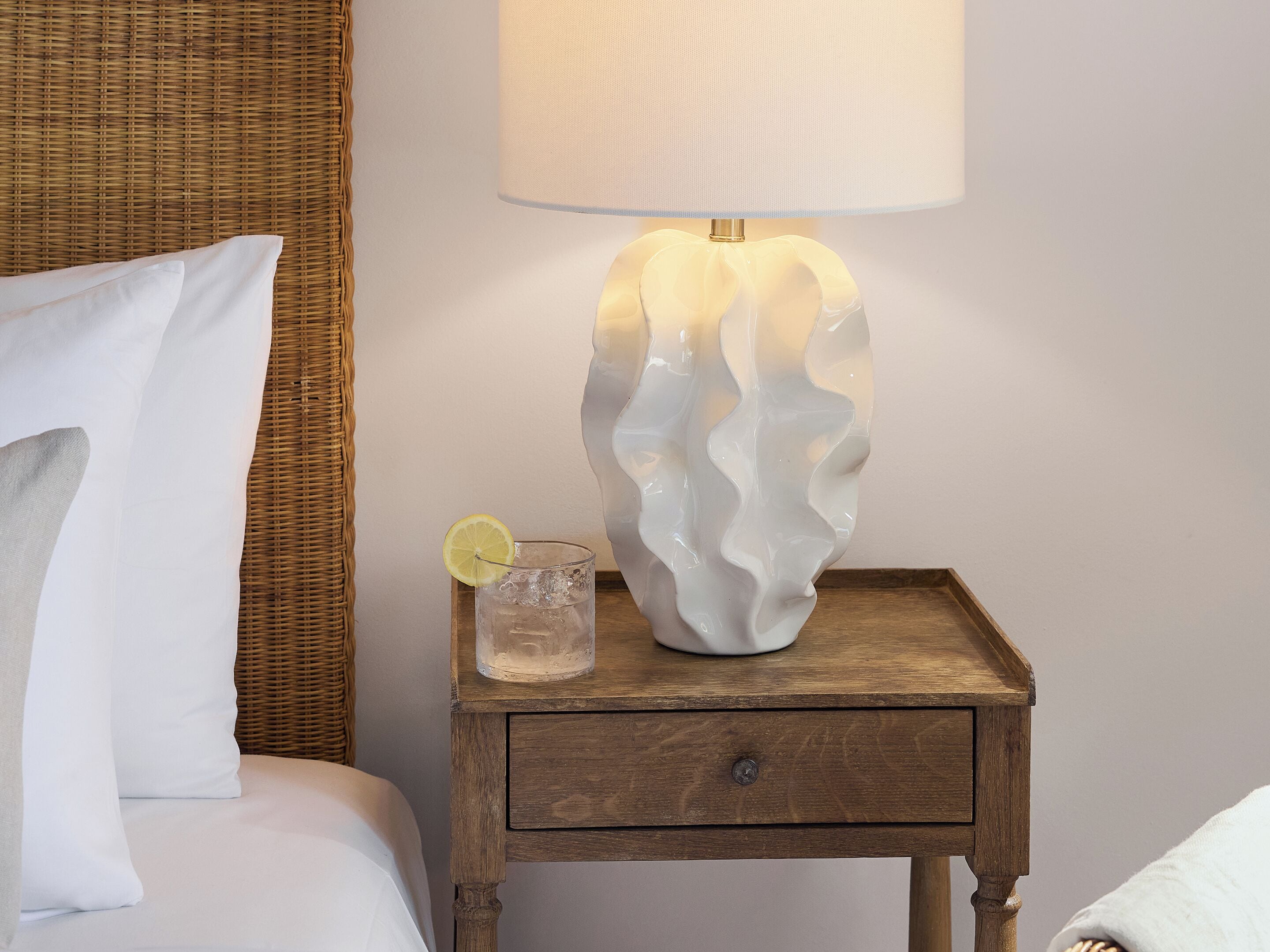 Ein Nachttisch mit Lampe, ein Glas Wasser mit Zitronenscheibe und ein Bett mit weißer Bettwäsche.