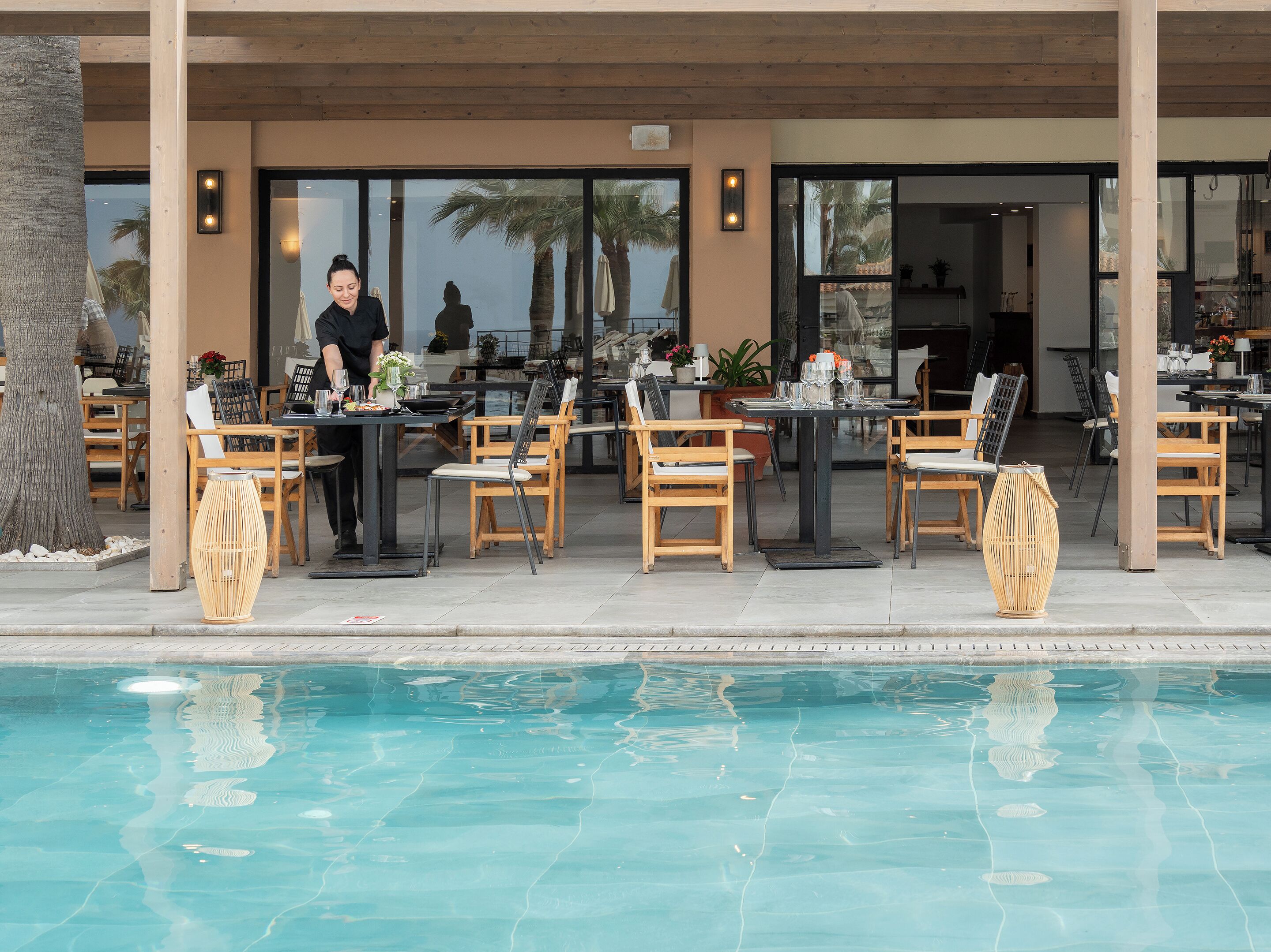 An outdoor dining area with tables and chairs, a pool in the foreground, and a view of the pool area through large windows.