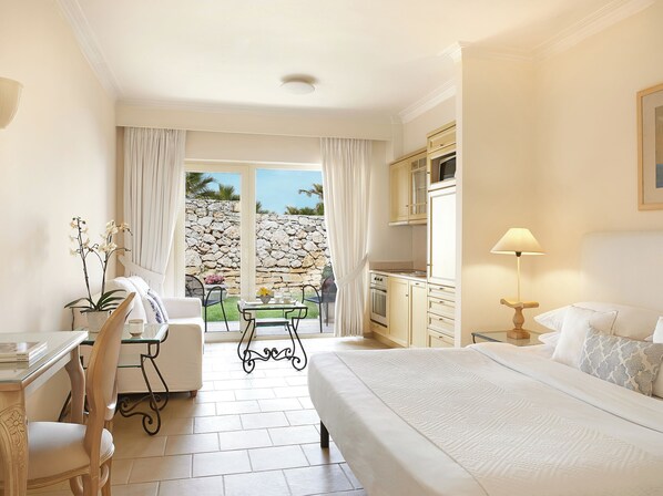 A hotel room with a bed, a desk, a chair, a table with a vase of flowers, a lamp, and a view of a stone wall and greenery outside.