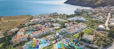 Vista aérea de un complejo turístico costero con varias edificaciones, un parque acuático grande y vistas al mar y a las montañas.