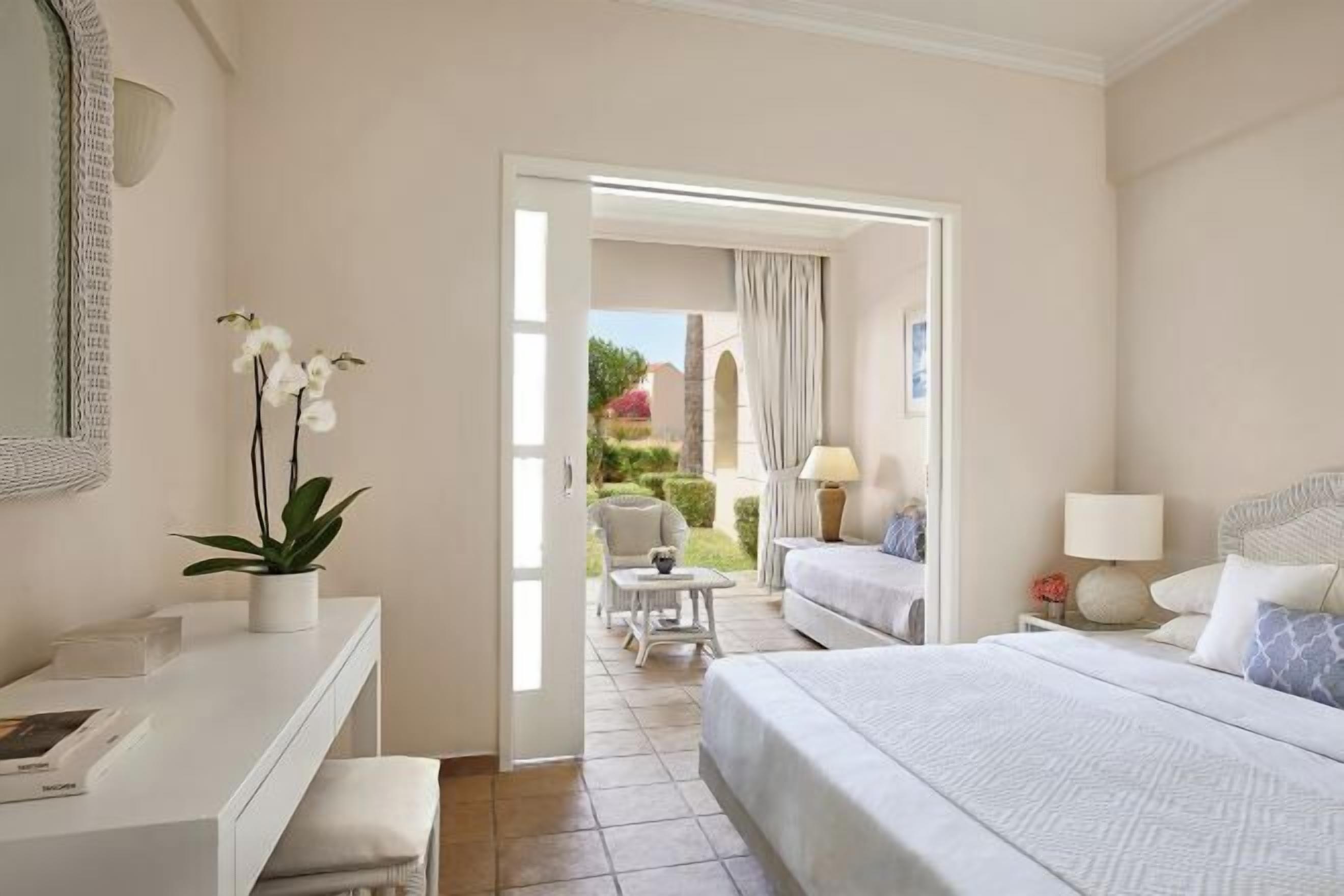 A hotel room with a bed, bedside table, lamp, and a view of the garden through an open door.