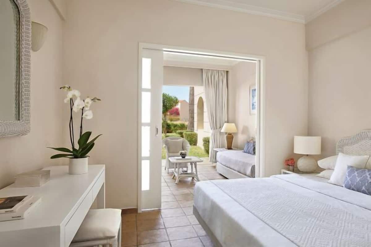 A hotel room with a bed, bedside table, lamp, and a view of the garden through an open door.