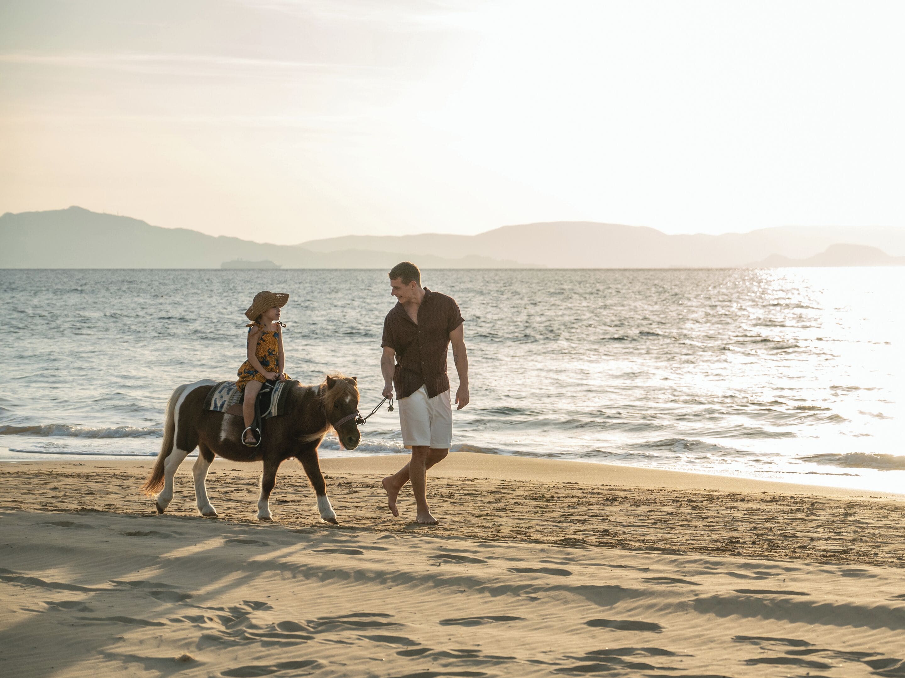 Escena de playa con una persona guiando un caballo y un niño montándolo.
