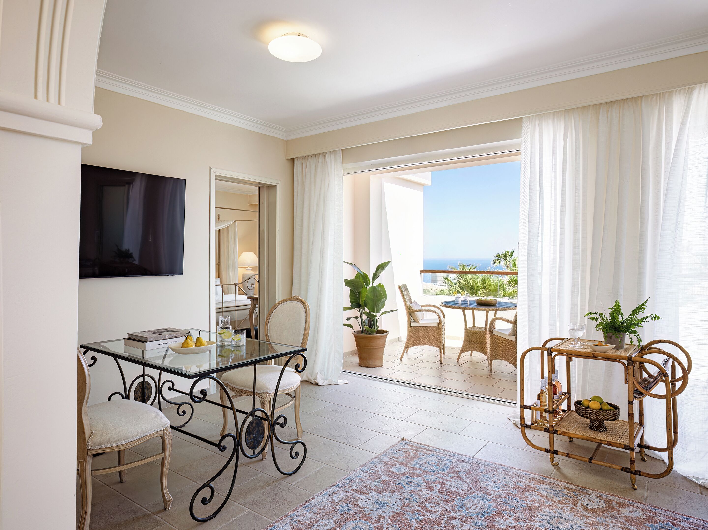 Habitación de hotel con balcón, comedor con mesa de vidrio, sillas de mimbre y una pequeña mesita con una planta.