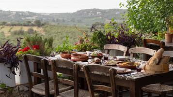 A rustic outdoor dining table with terracotta pots, wicker chairs, and a view of a hilly landscape.