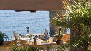 An outdoor seating area with a table and chairs, overlooking a body of water, with potted plants and a wooden ceiling structure.