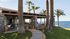 A coastal outdoor dining area with palm trees, stone walls, and a view of the sea.