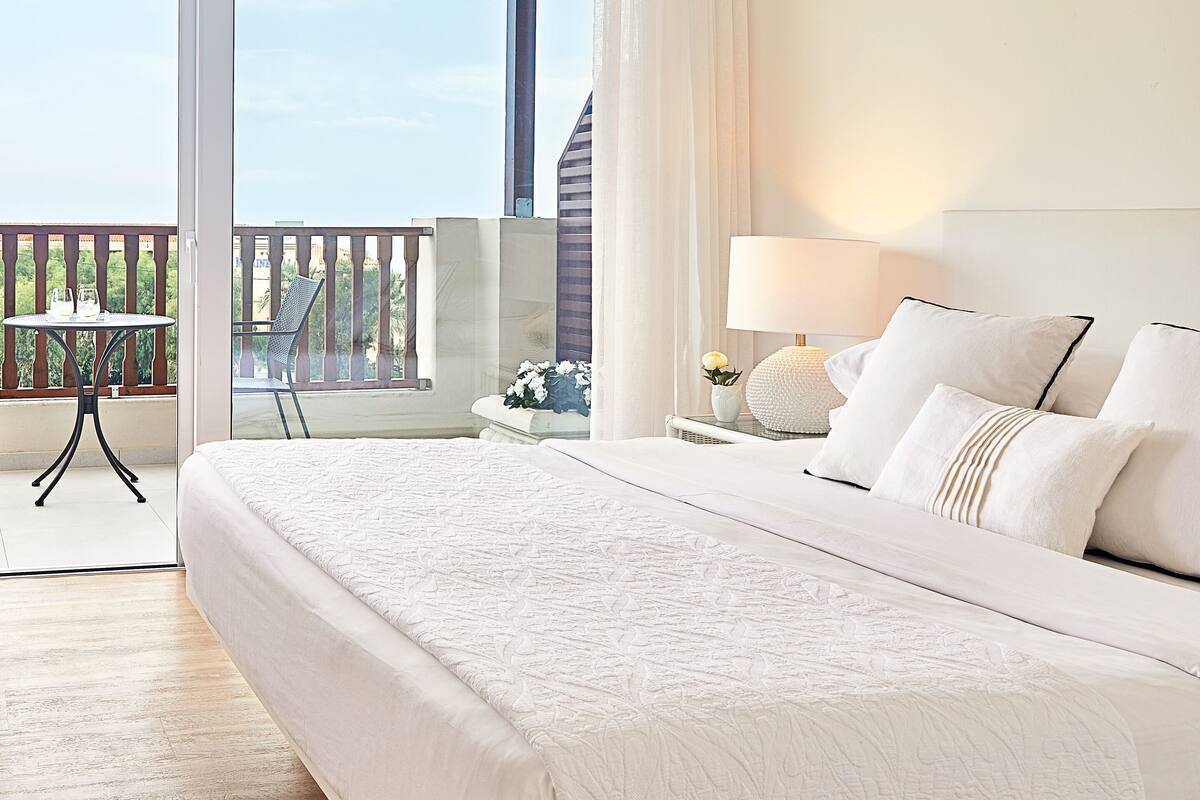 A hotel room with a large bed, a small table, a chair, a nightstand, and a balcony with a view.