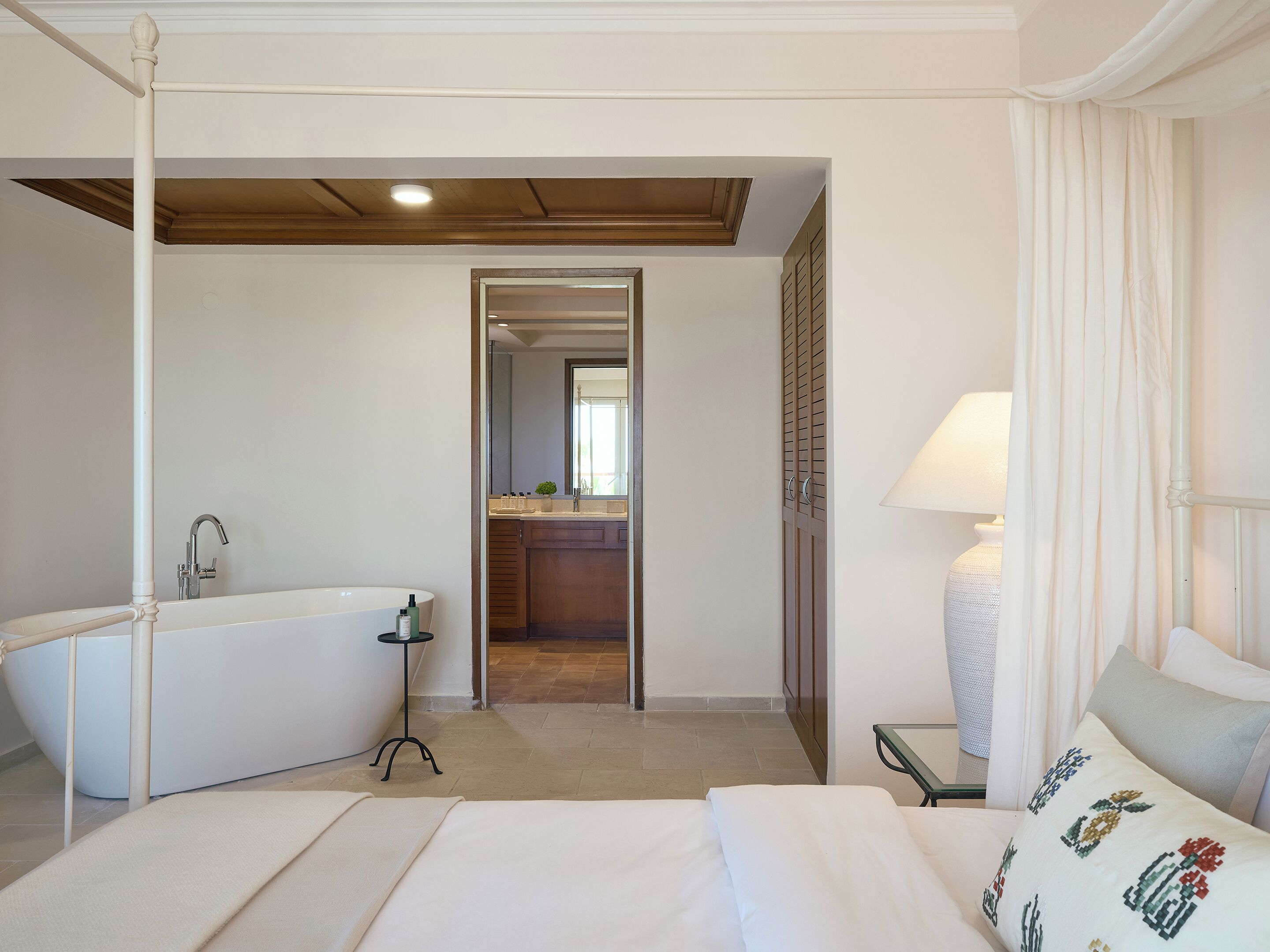 A hotel room with a large bed, a separate bathroom with a bathtub, a small table with a lamp, and a view of the hallway.