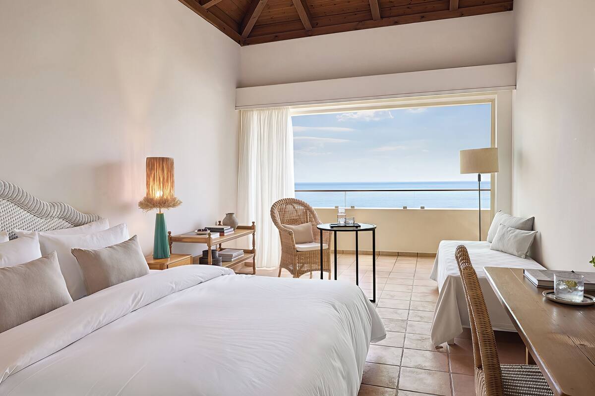 A hotel room with a large bed, a dining table, a wicker chair, and a view of the ocean.