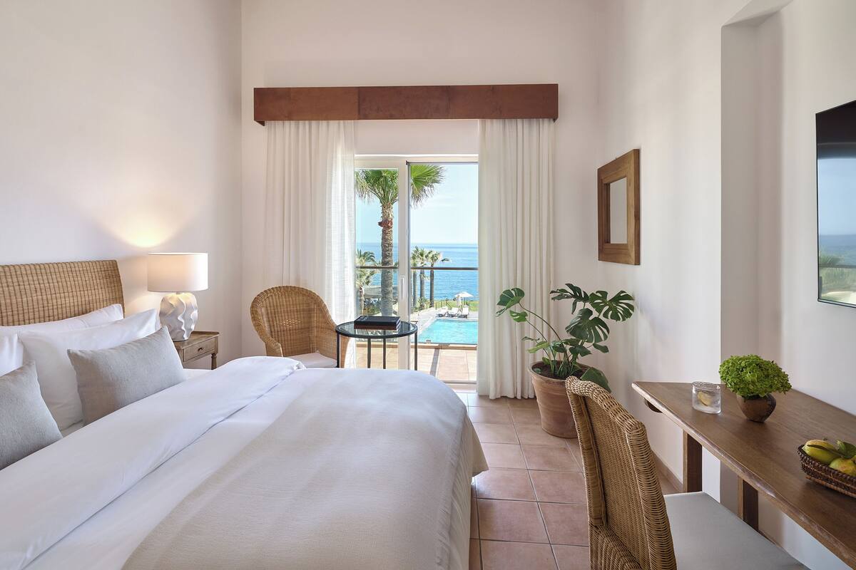 A hotel room with a bed, a table with a plant, a chair, and a view of the ocean through a large window.