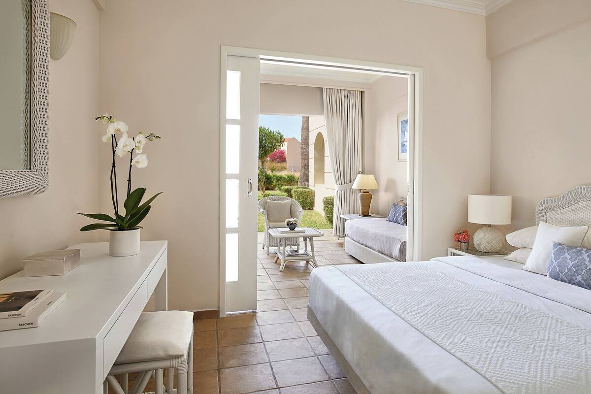 A hotel room with a bed, a desk, a chair, a small table, a lamp, and a potted plant.