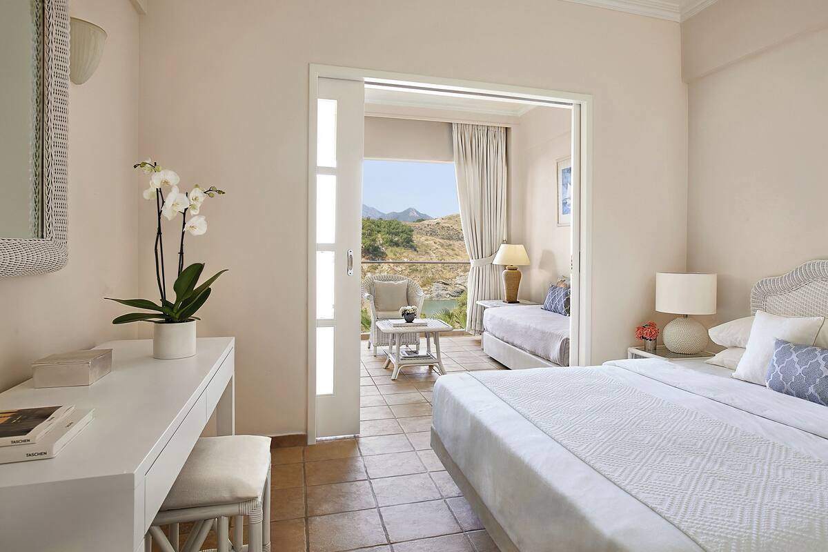 A hotel room with a bed, a desk, a chair, a small table, a lamp, and a view of the outdoors.