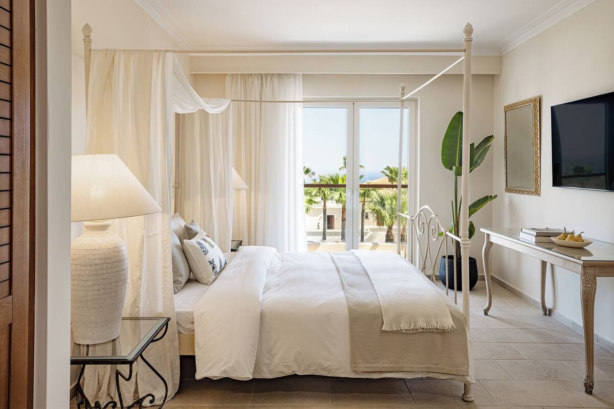 A bedroom with a bed, a bedside table, a lamp, a TV, and a view of the outdoors.