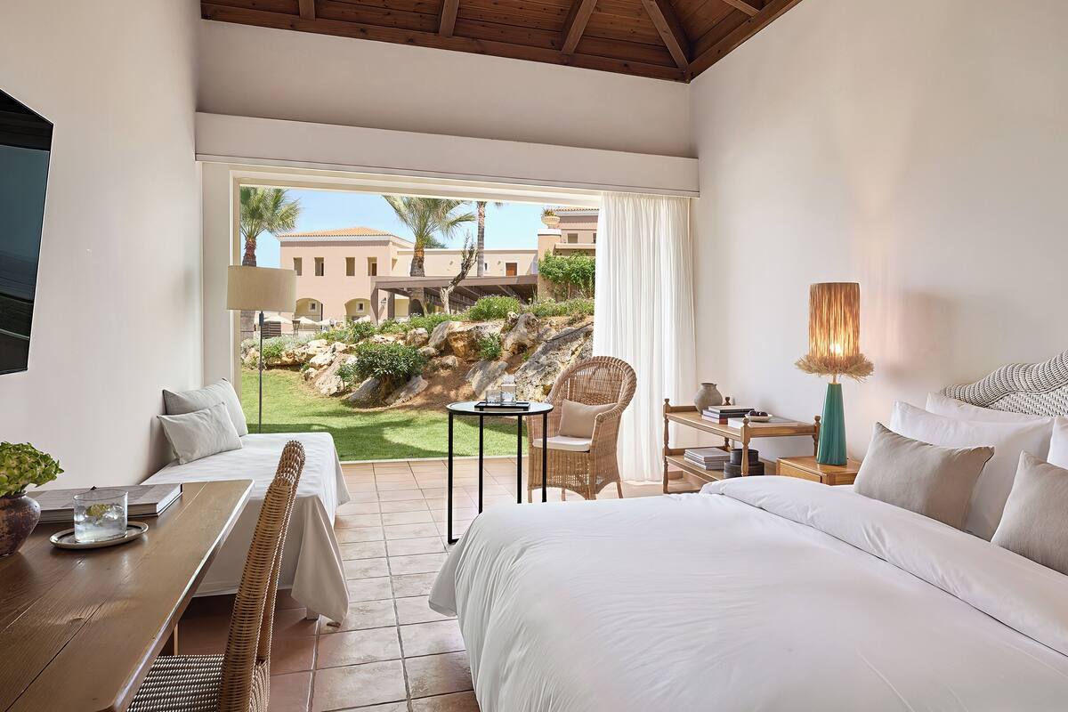 A hotel room with a bed, a desk, a chair, a lamp, and a view of the outside.