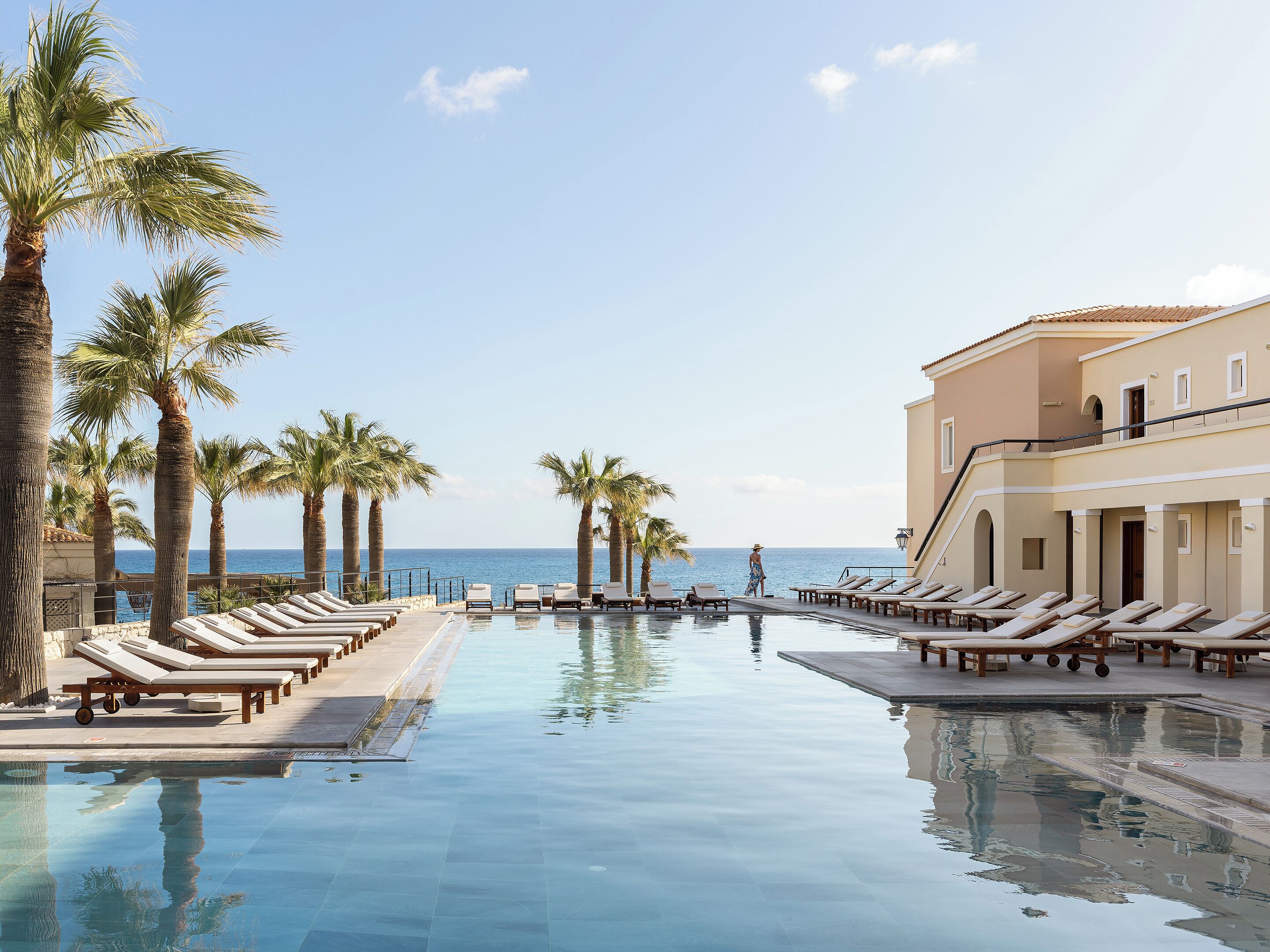 A pool area with lounge chairs and palm trees, overlooking the sea.
