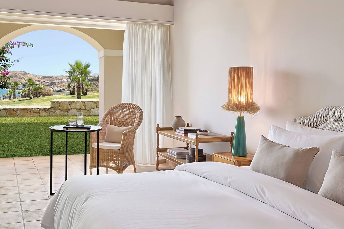 A hotel room with a bed, a wicker chair, a small table, and a view of a landscaped area.