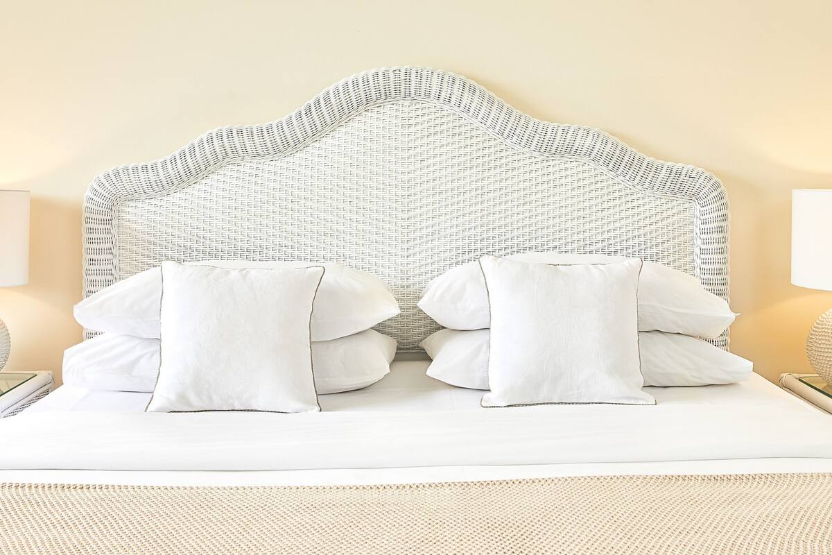 A neatly made bed with white linens and pillows, a woven headboard, and a textured rug.