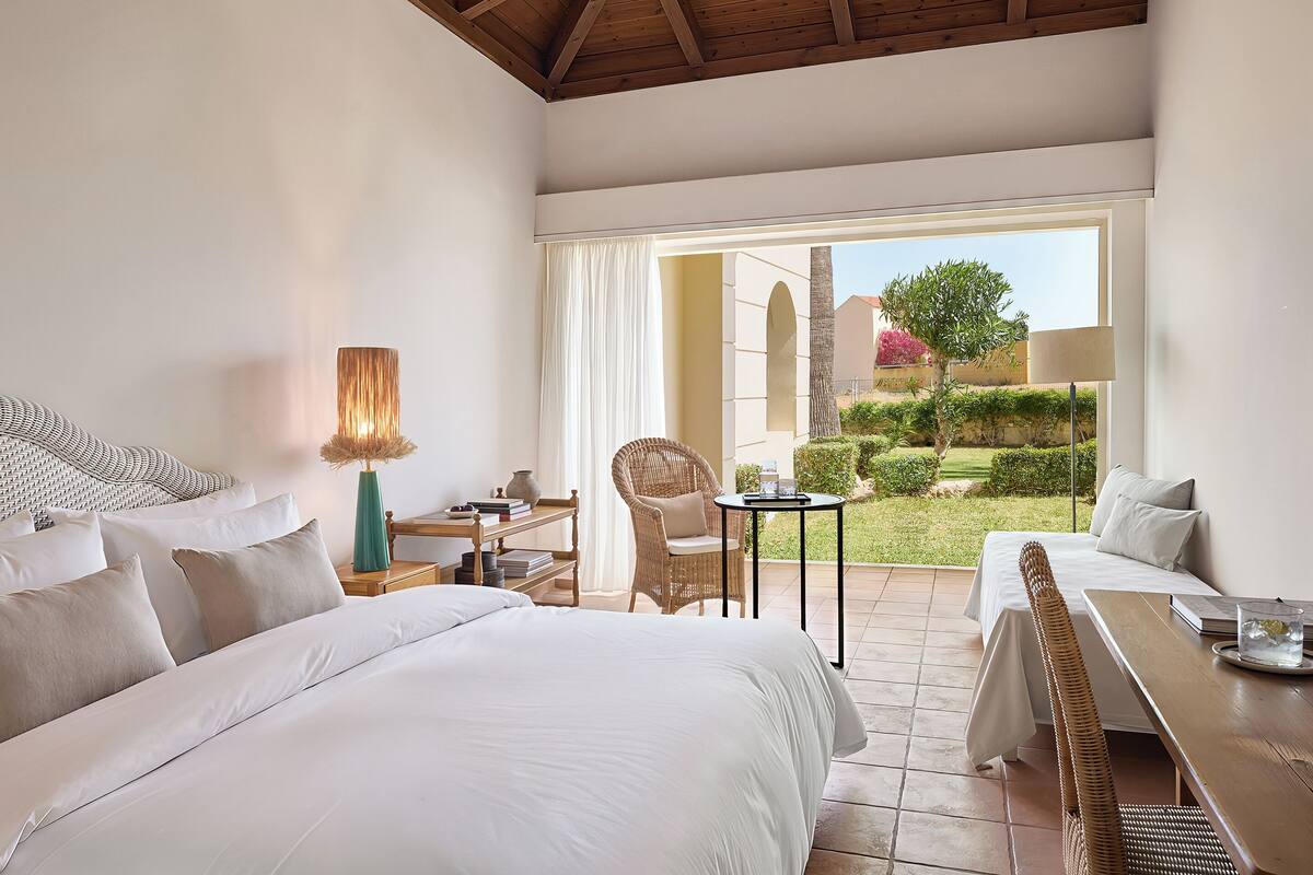 A bedroom with a large bed, a small table, a chair, and a view of a garden.