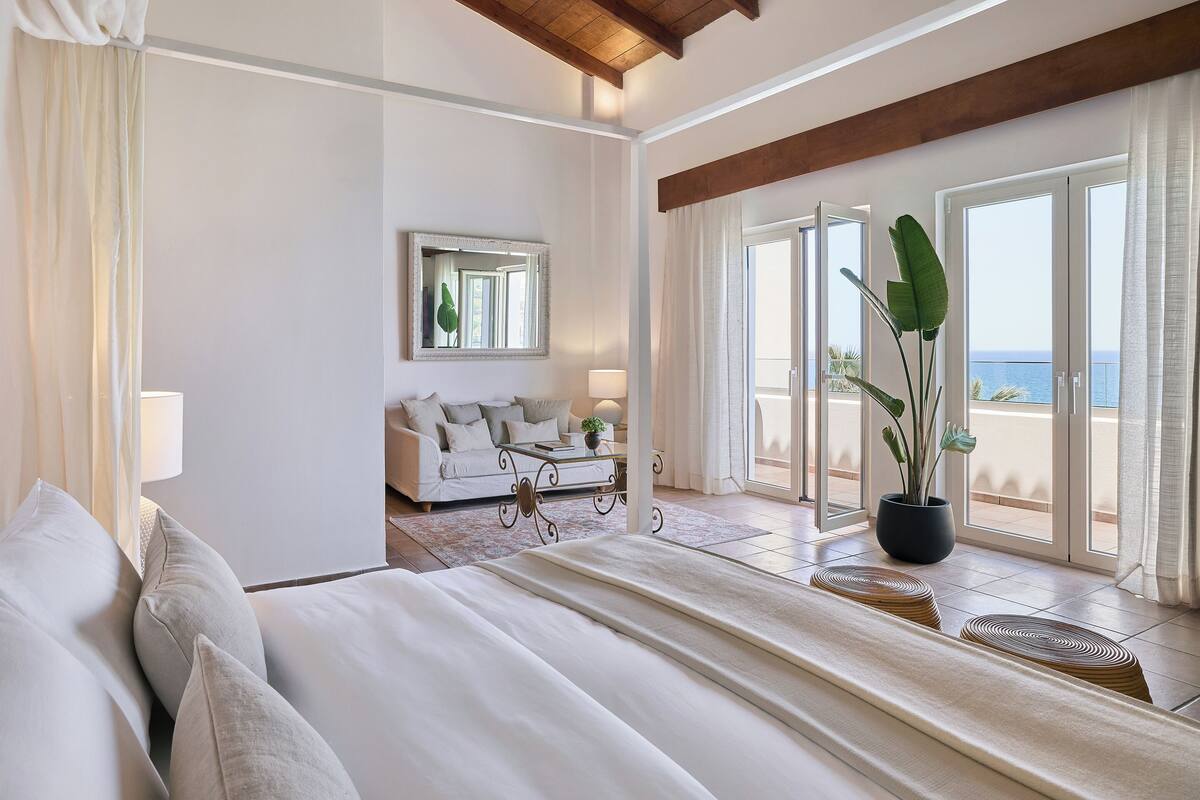 A bedroom with a bed, a sofa, a large mirror, a potted plant, and a view of the sea through a sliding door.