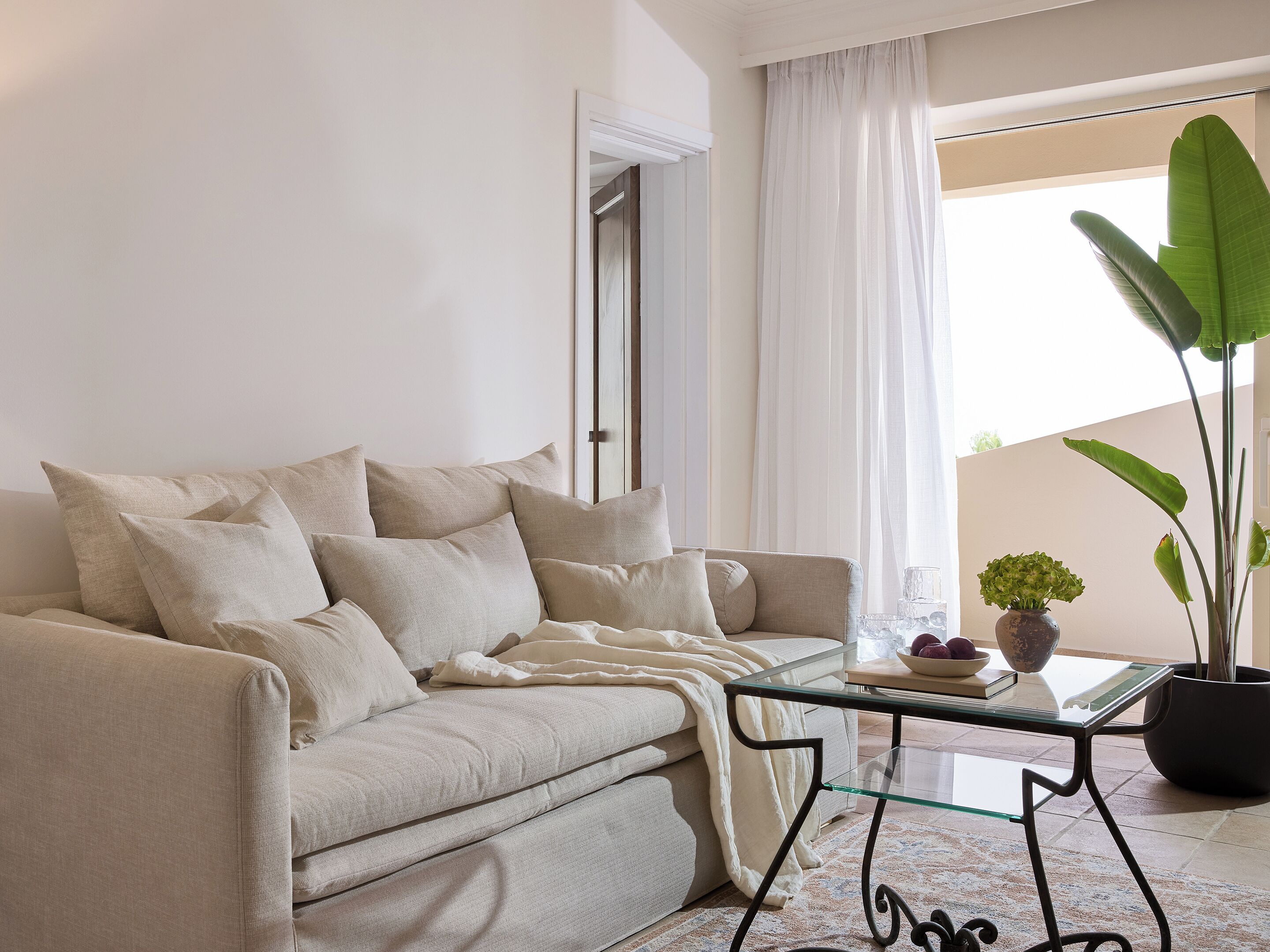 A living room with a beige sofa, a glass coffee table, a potted plant, and a view of the outdoors.