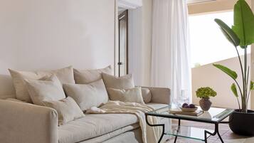 A living room with a beige sofa, a glass coffee table, a potted plant, and a view of the outdoors.