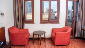 A room with two red sofas, a small wooden table, and a view of buildings through large windows. - Hotel Ambassadeur (Nairobi)