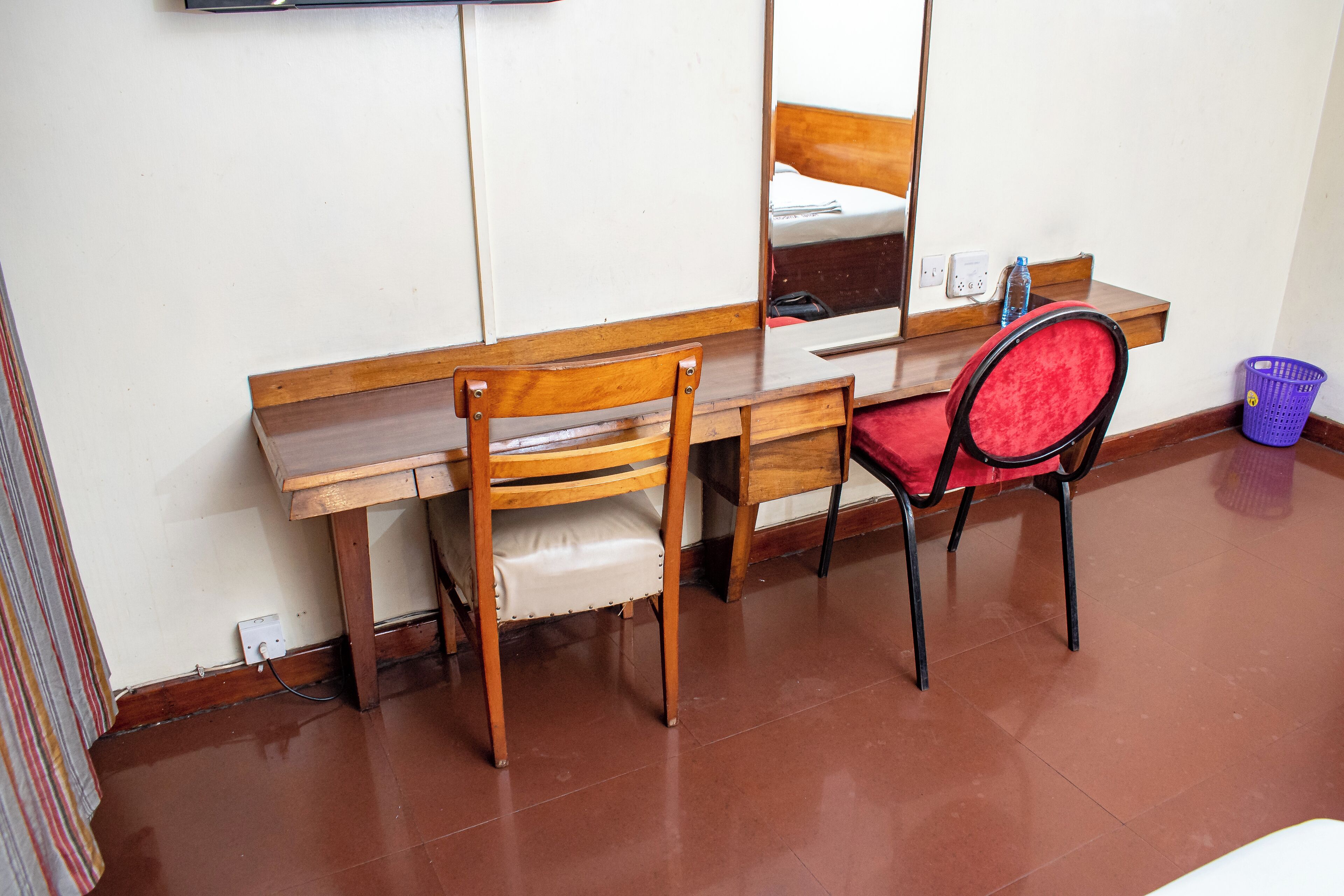Een houten bureau met een spiegel, twee stoelen, een rode kussen, een paarse wasmand en een witte stopcontact.