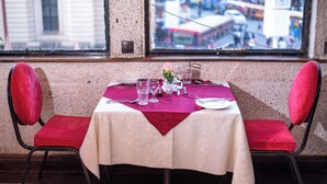 A dining table set with a red tablecloth, two red cushioned chairs, and a window view of a busy street. - Hotel Ambassadeur (Nairobi)