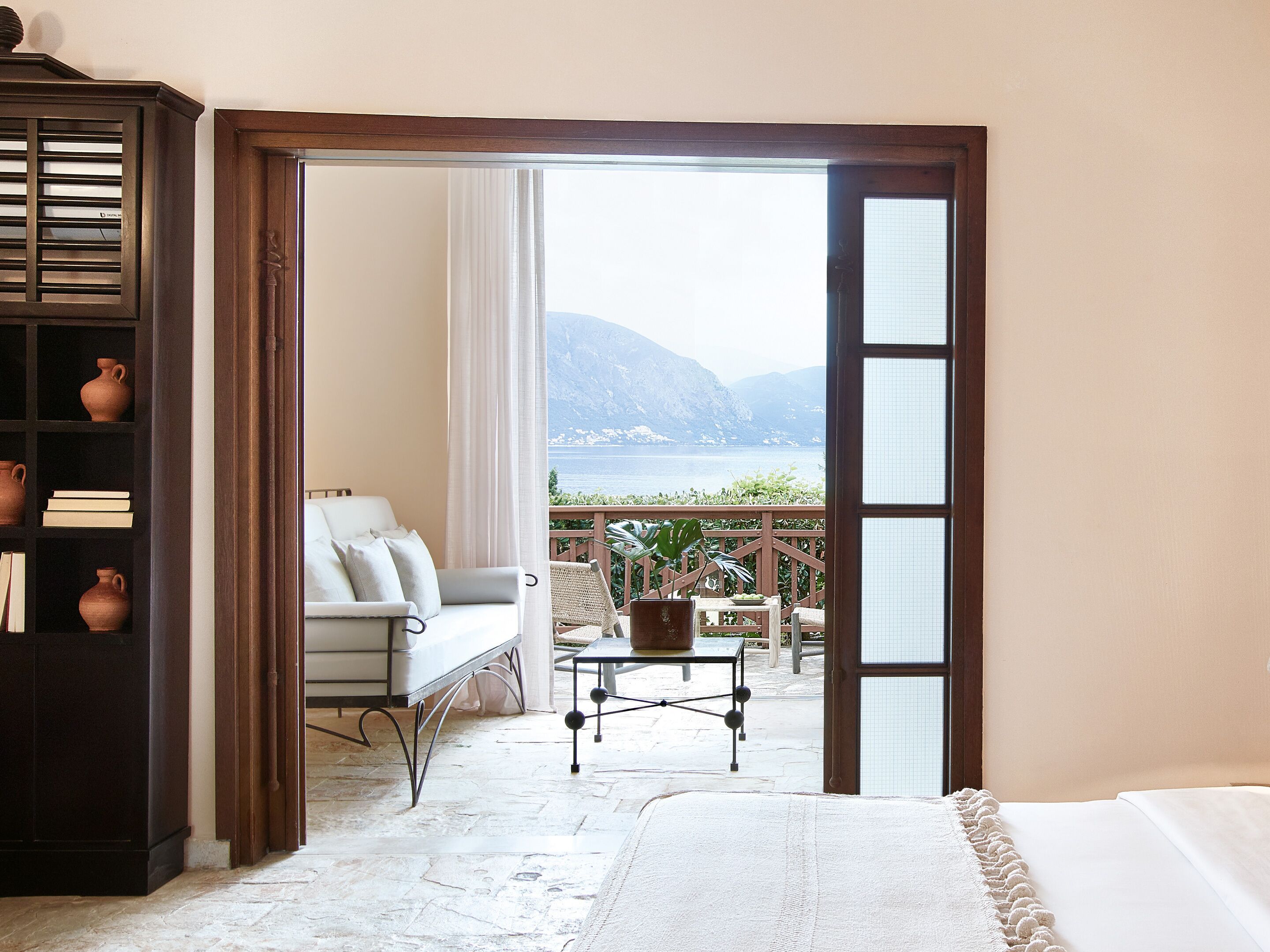 Una habitación con cama, sofá, una mesita y una estantería. Se ve el mar y las montañas a través de una puerta abierta.
