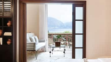 Una habitación con cama, sofá, una mesita y una estantería. Se ve el mar y las montañas a través de una puerta abierta.