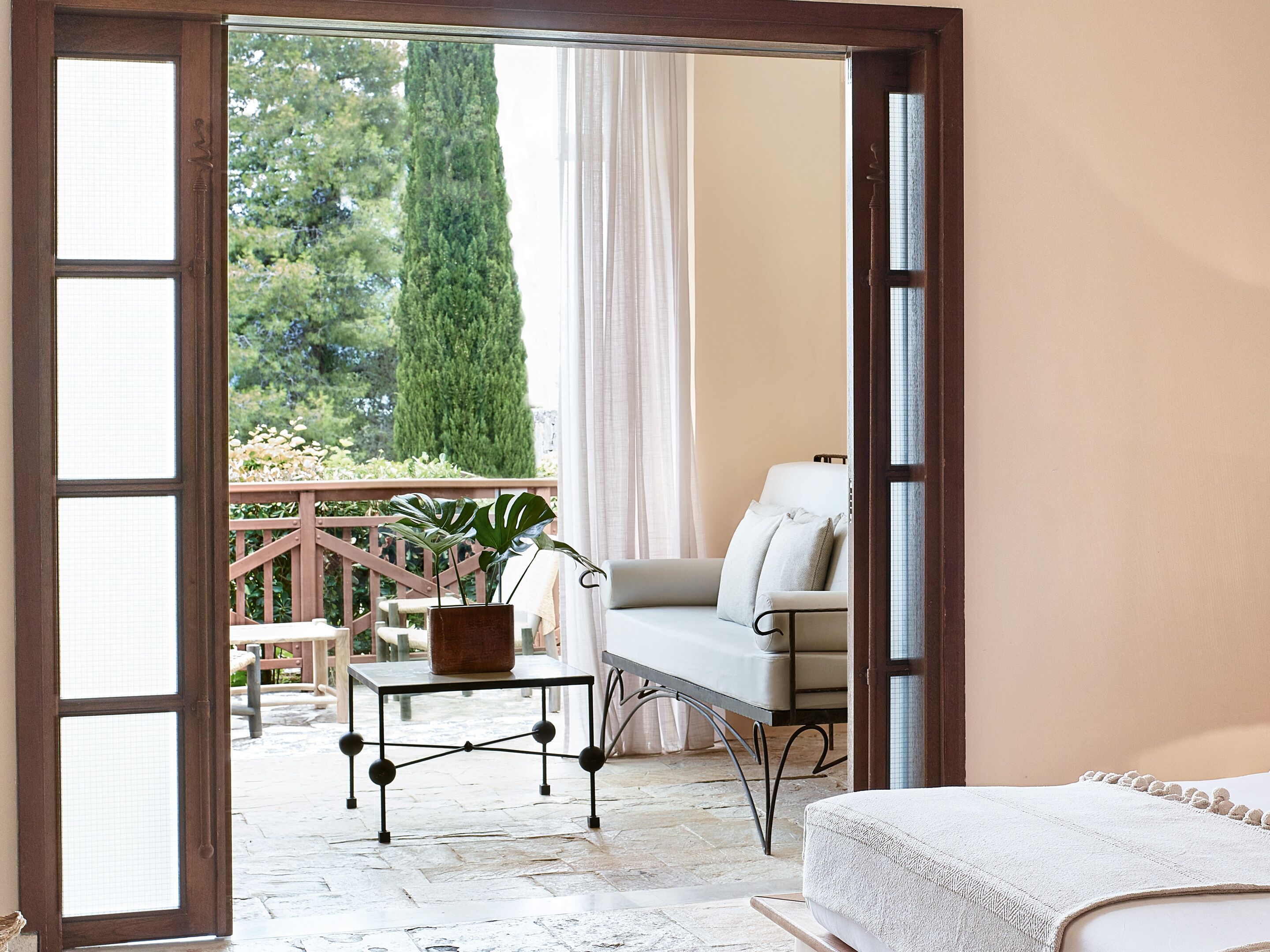 A room with a bed, a table with a potted plant, a chair, and a view of a garden through a glass door.
