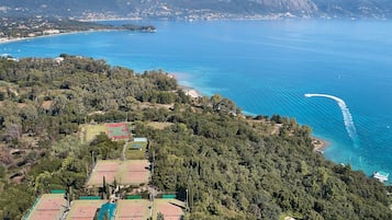 Vista aérea de una zona costera con un complejo de canchas de tenis rodeado de densa vegetación, una playa y montañas al fondo.