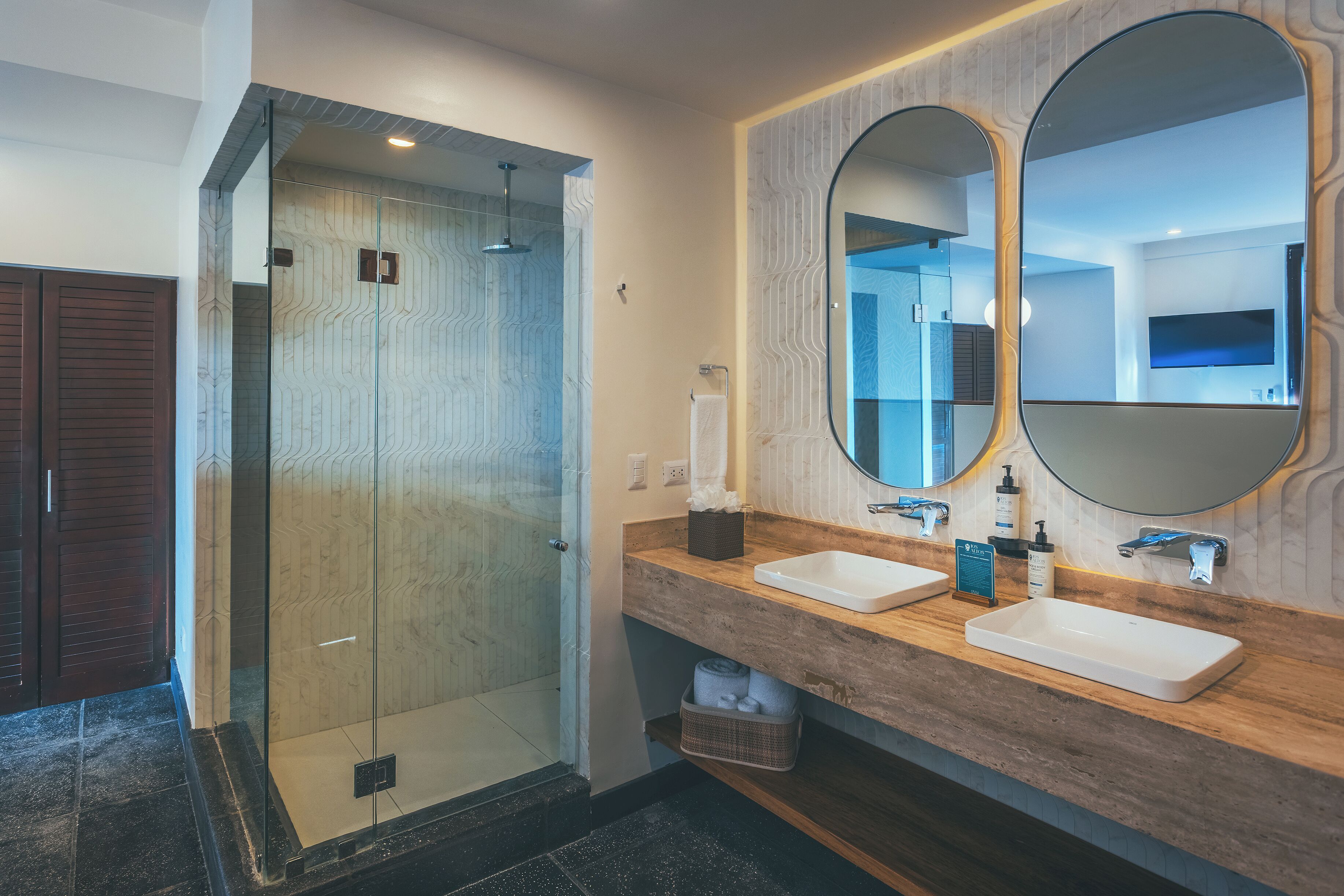 Un baño moderno con ducha envuelta en vidrio, dos lavamanos con espejos y un mueble de tocador de madera.