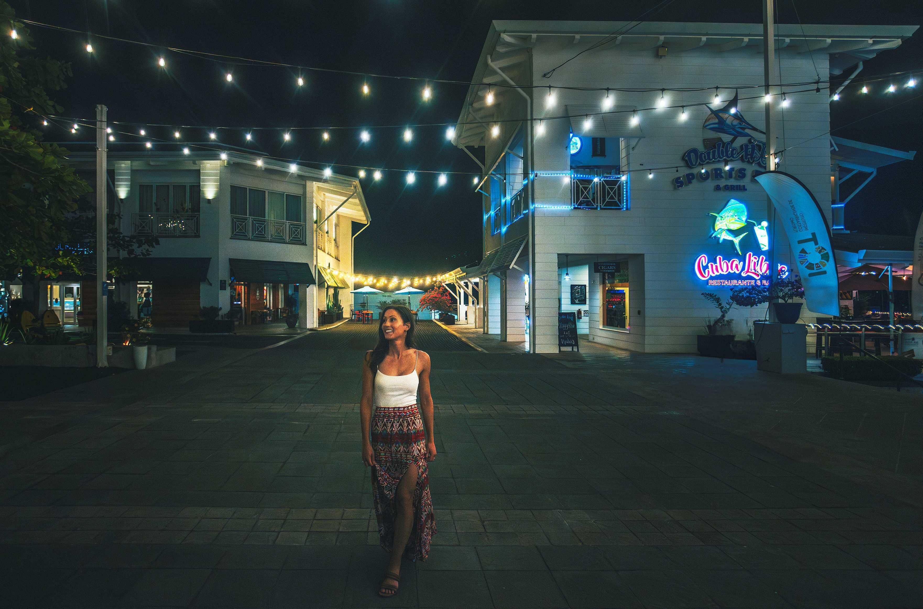 Una mujer caminando sobre una superficie pavimentada con luces colgantes. En el fondo, se ven edificios con letreros que dicen 'Cuba Lita' y 'Violetta's Sports Bar & Grill'.