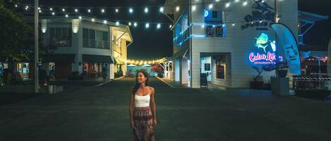 Una mujer caminando sobre una superficie pavimentada con luces colgantes. En el fondo, se ven edificios con letreros que dicen 'Cuba Lita' y 'Violetta's Sports Bar & Grill'.