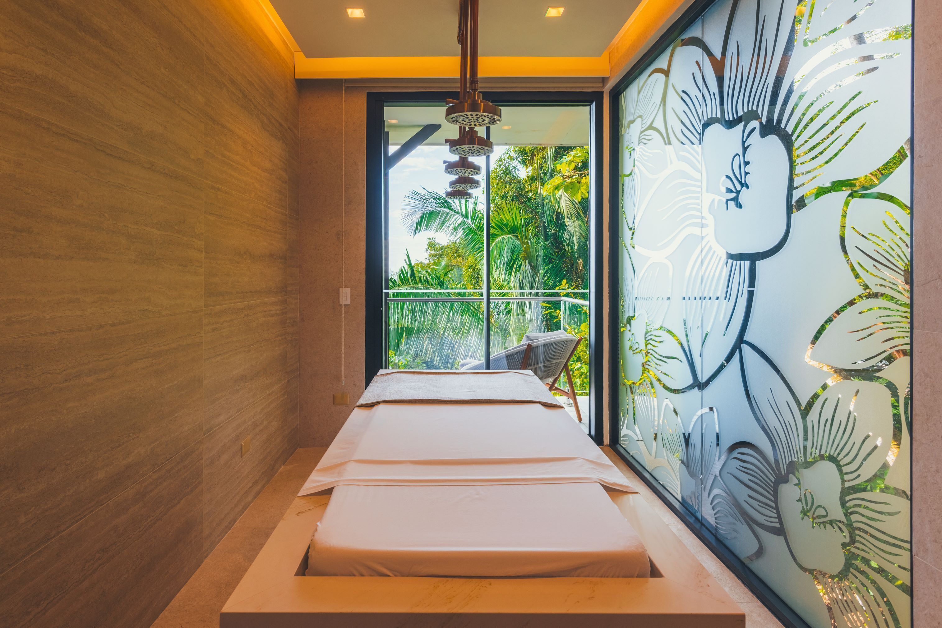 Ein Spa-Raum mit Massagetisch, einer Glaswand mit tropischem Wandbild und Blick auf Grünflächen.
