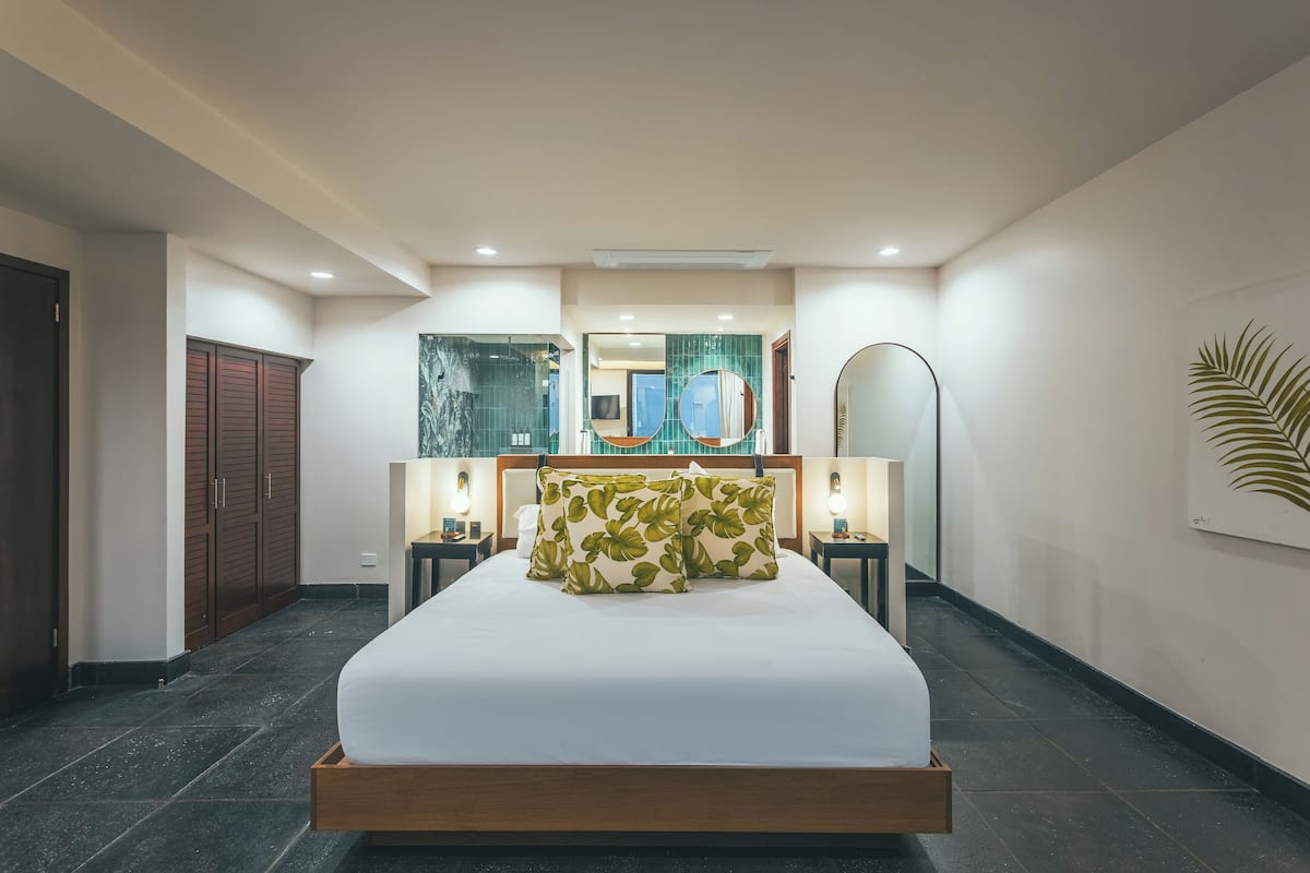 Une chambre d’hôtel moderne avec un grand lit, une tête de lit en bois et un miroir au-dessus. On y trouve deux tables de chevet avec des lampes, une armoire et une grande fenêtre donnant sur un beau paysage.
